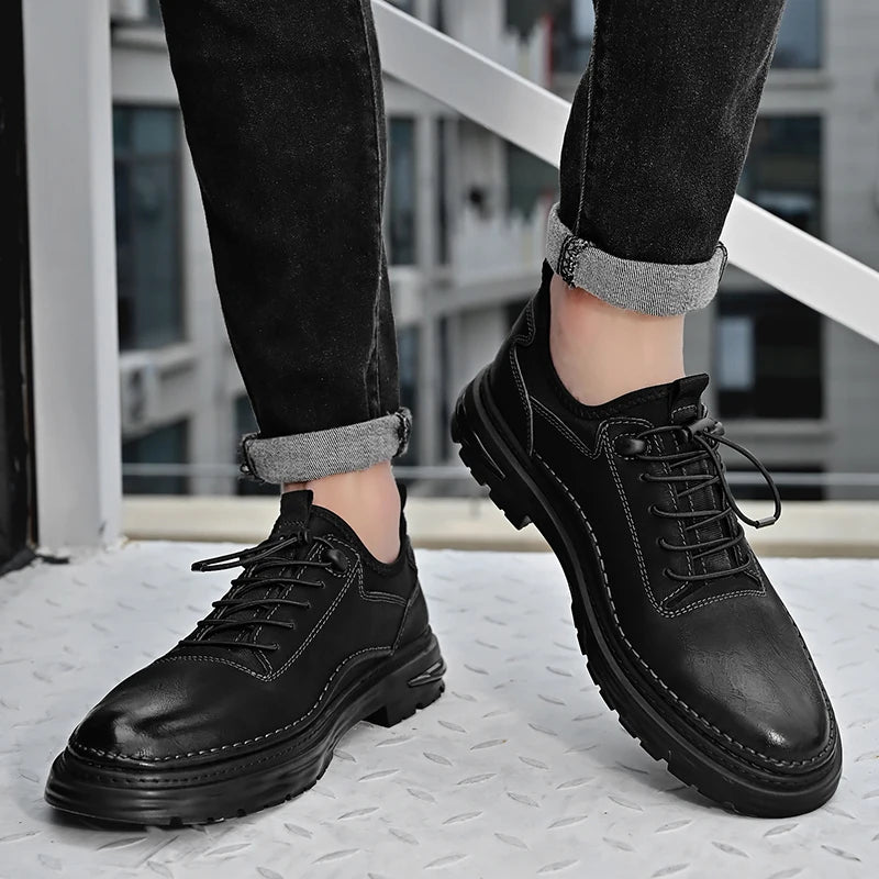 Black leather shoes worn with rolled-up jeans on a textured floor.