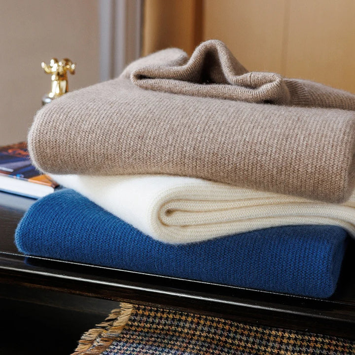 Stack of folded sweaters in beige, white, and blue on a surface.
