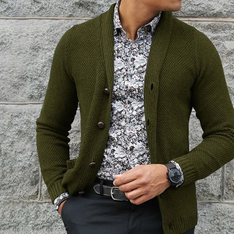 Person wearing a green cardigan over a patterned shirt against a stone wall.