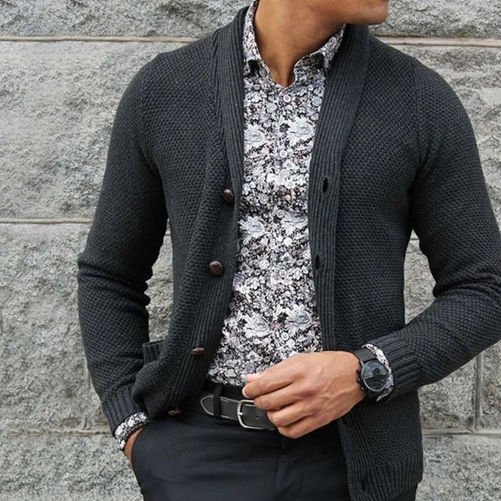 Person wearing a dark gray cardigan over a patterned shirt against a stone wall.