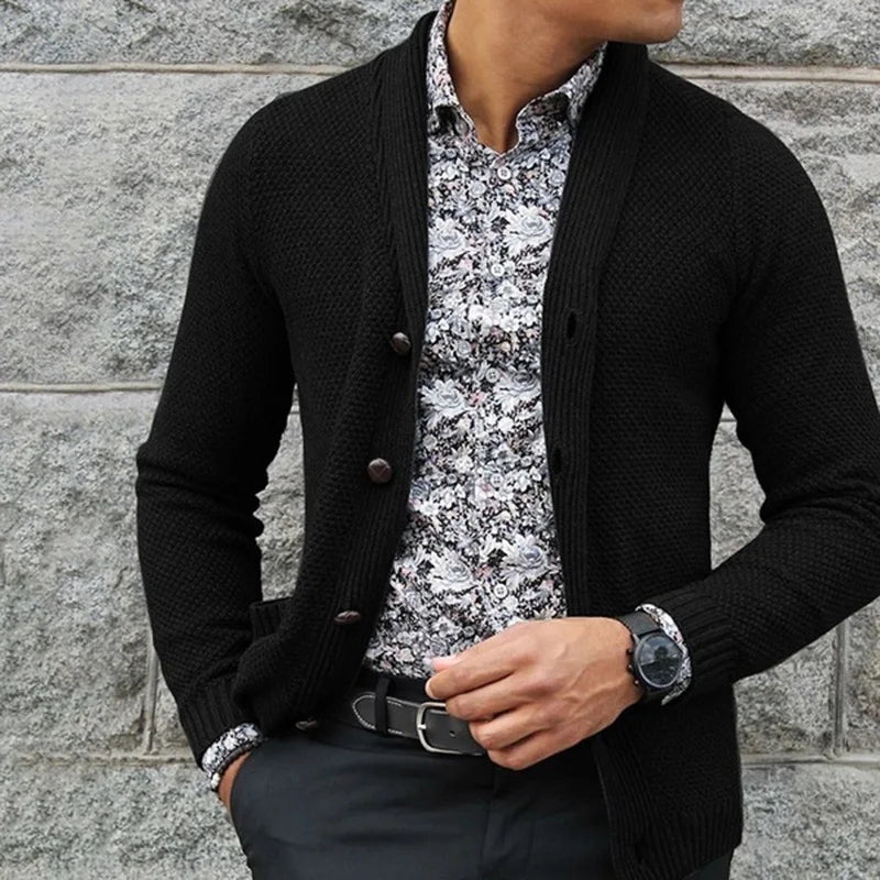 Person wearing a black cardigan over a patterned shirt against a stone wall.
