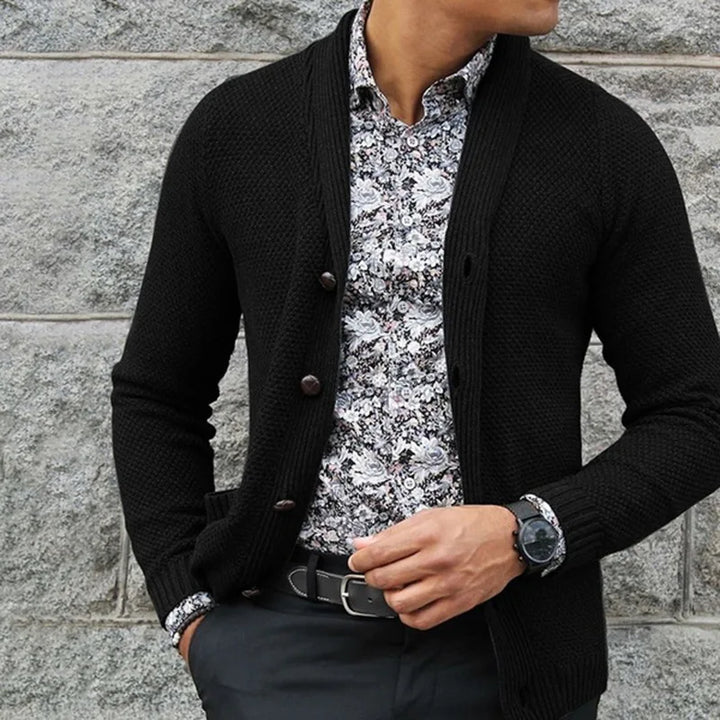 Person wearing a black cardigan over a patterned shirt against a stone wall.