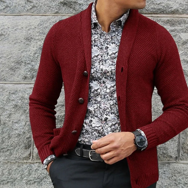 Person wearing a red cardigan over a patterned shirt against a stone wall.