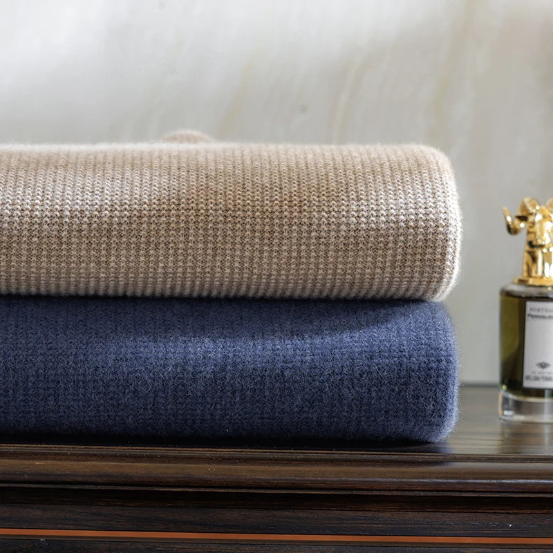 Two folded blankets, one beige and one blue, stacked on a wooden surface with a bottle of perfume in the background.