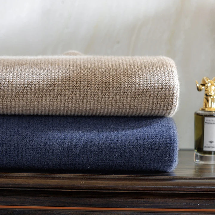 Two folded blankets, one beige and one blue, stacked on a wooden surface with a bottle of perfume in the background.