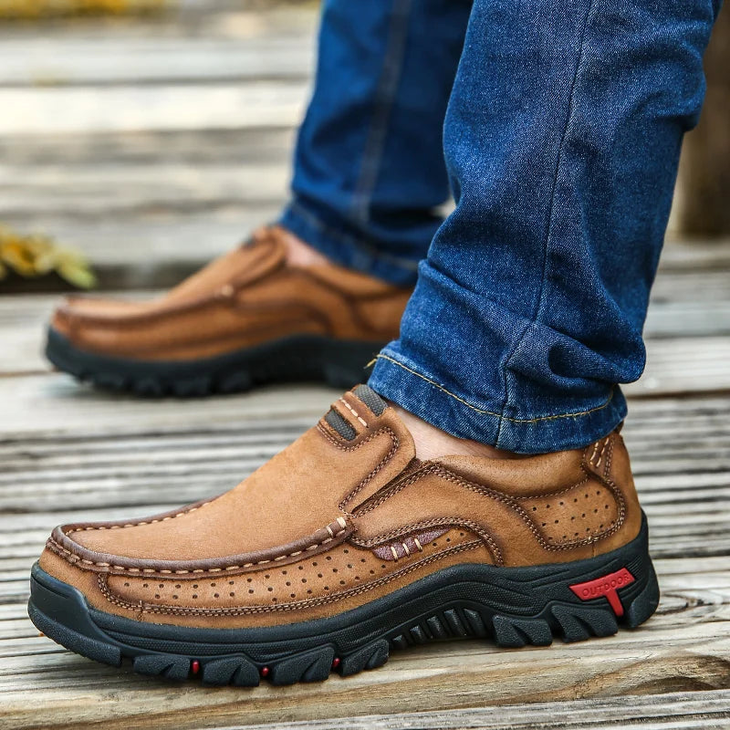 Brown leather shoes with black soles worn with blue jeans on a wooden floor.