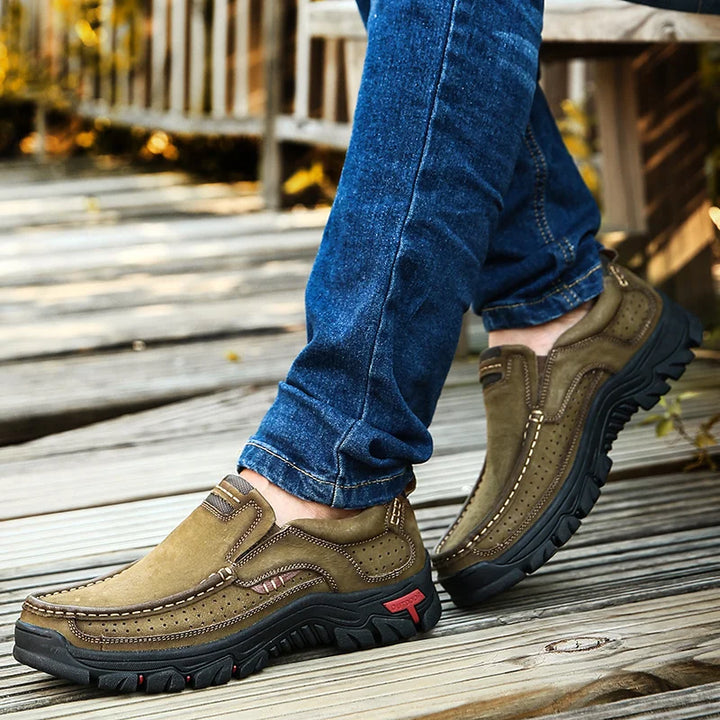 Brown leather shoes worn with blue jeans on a wooden floor.