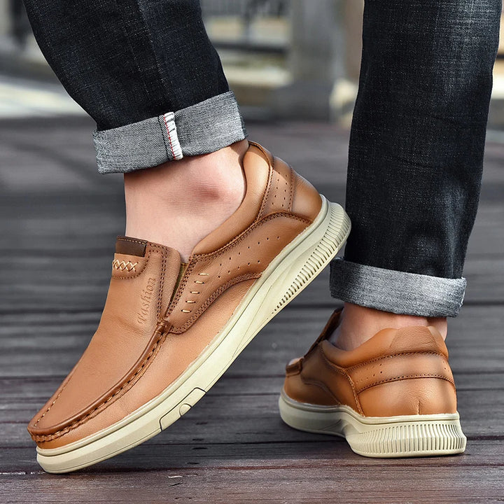 Brown leather shoes worn with rolled-up jeans on a wooden floor.