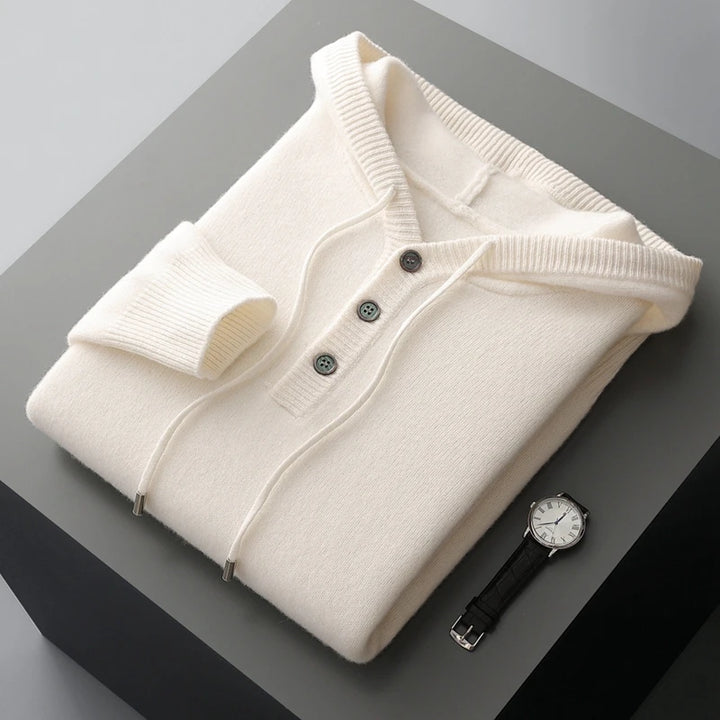 Folded cream-colored sweater with black buttons on a gray surface, accompanied by a black watch.