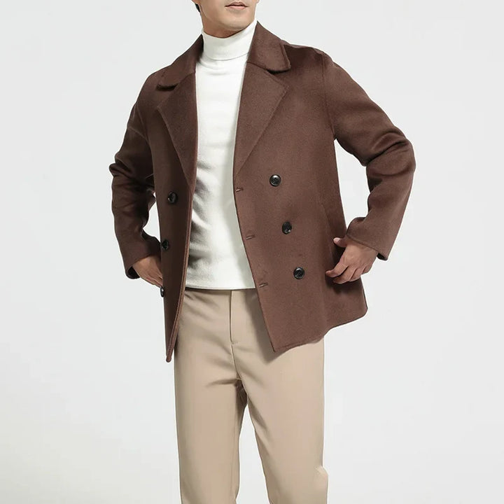 Man wearing a brown coat over a white turtleneck and beige pants on a light gray background