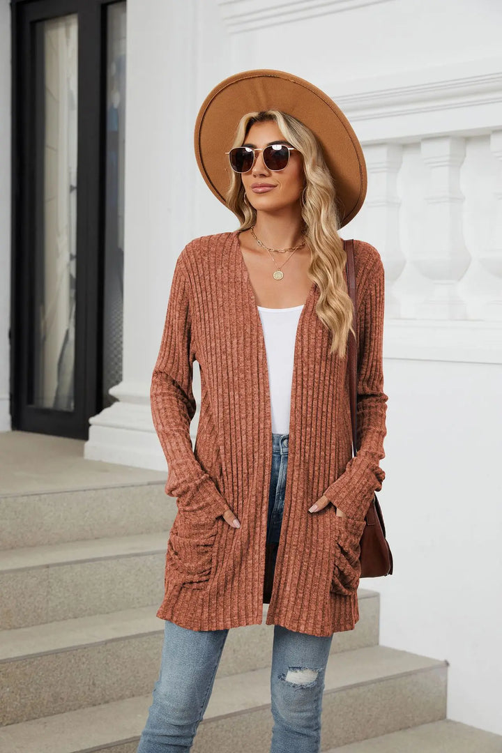 Woman wearing a brown cardigan, white top, jeans, and a brown hat outdoors.