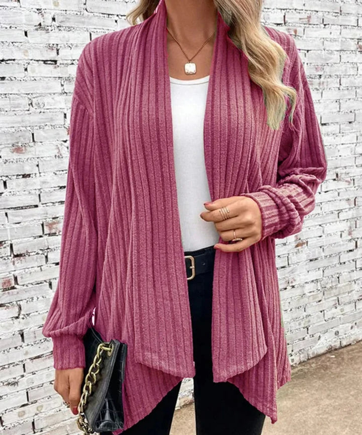 Person wearing a pink ribbed cardigan over a white top and black pants, standing against a white brick wall.