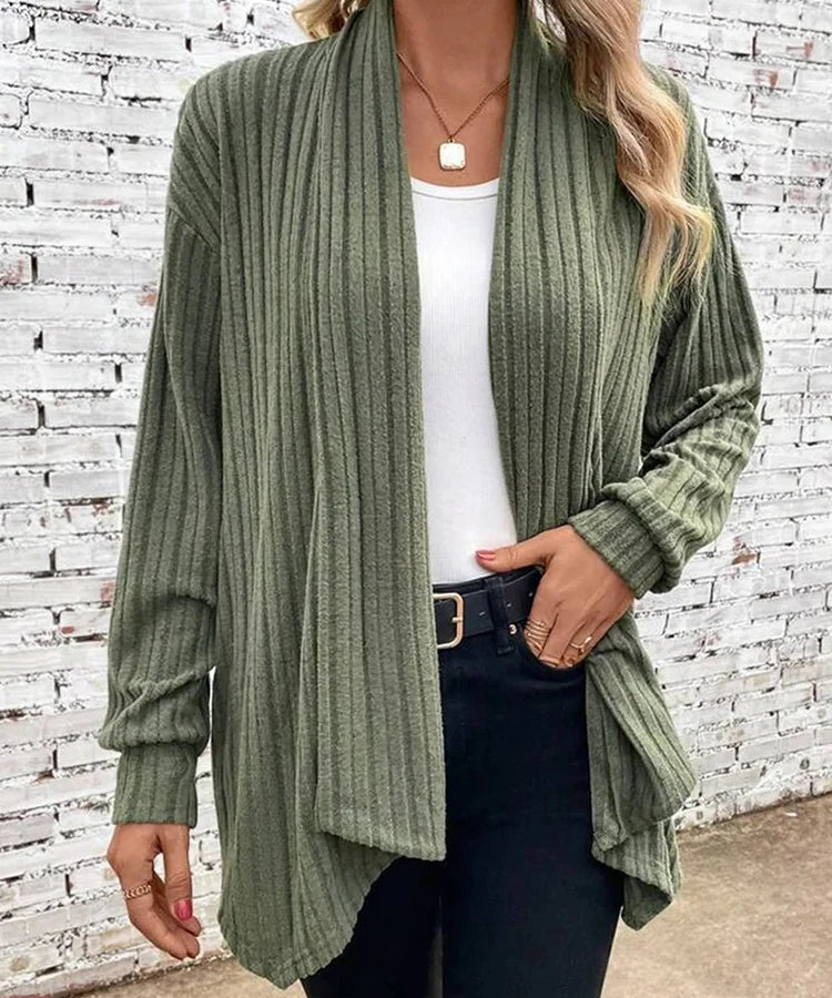 Woman wearing a green ribbed cardigan over a white top and black pants against a white brick wall.