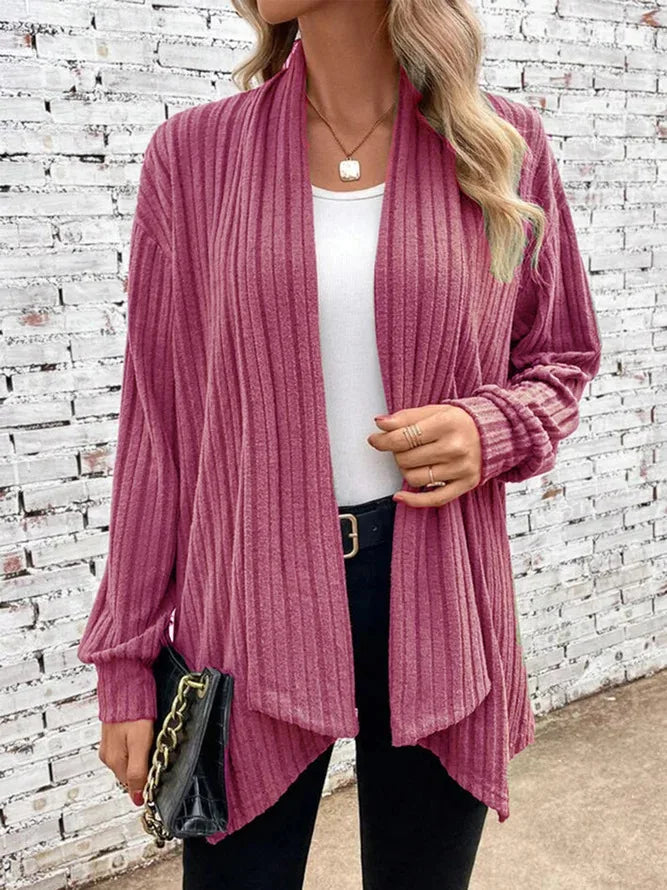 Person wearing a pink ribbed cardigan over a white top and black pants, standing against a white brick wall.