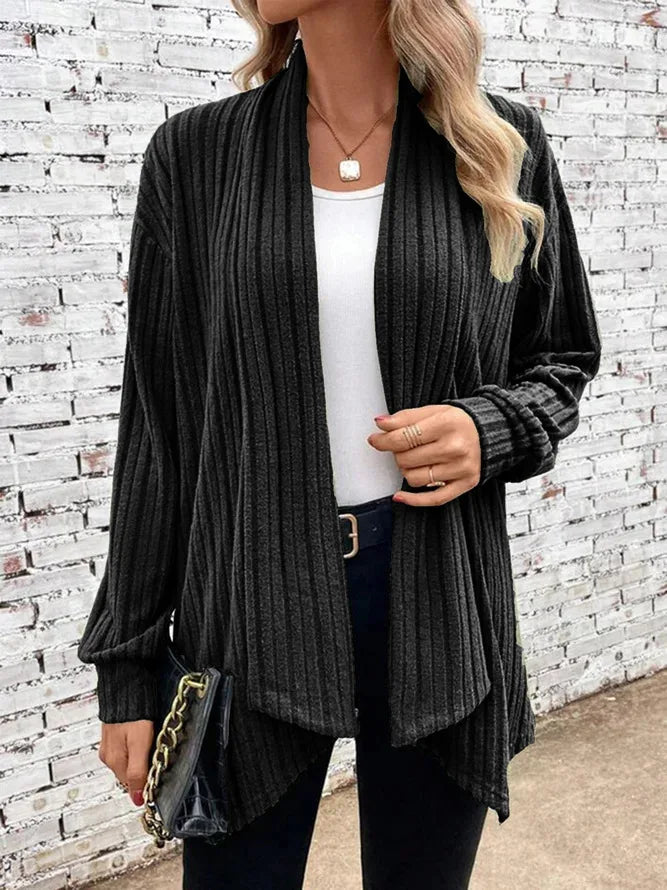 Woman wearing a dark ribbed cardigan over a white top and black pants, holding a black handbag against a white brick wall.