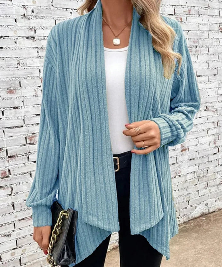 Woman wearing a light blue ribbed cardigan over a white top and black pants, standing against a white brick wall.