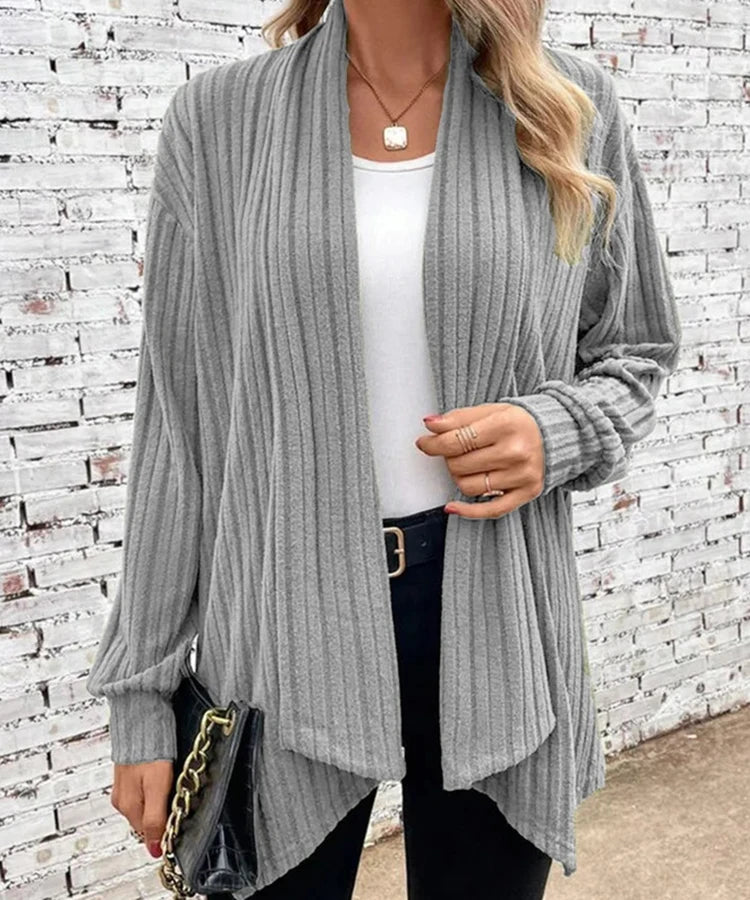 Person wearing a gray ribbed cardigan over a white top and black pants, standing against a white brick wall.