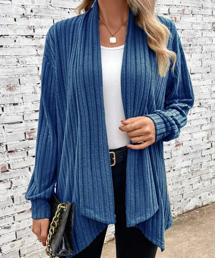 Woman wearing a blue ribbed cardigan over a white top and black pants, standing against a white brick wall.