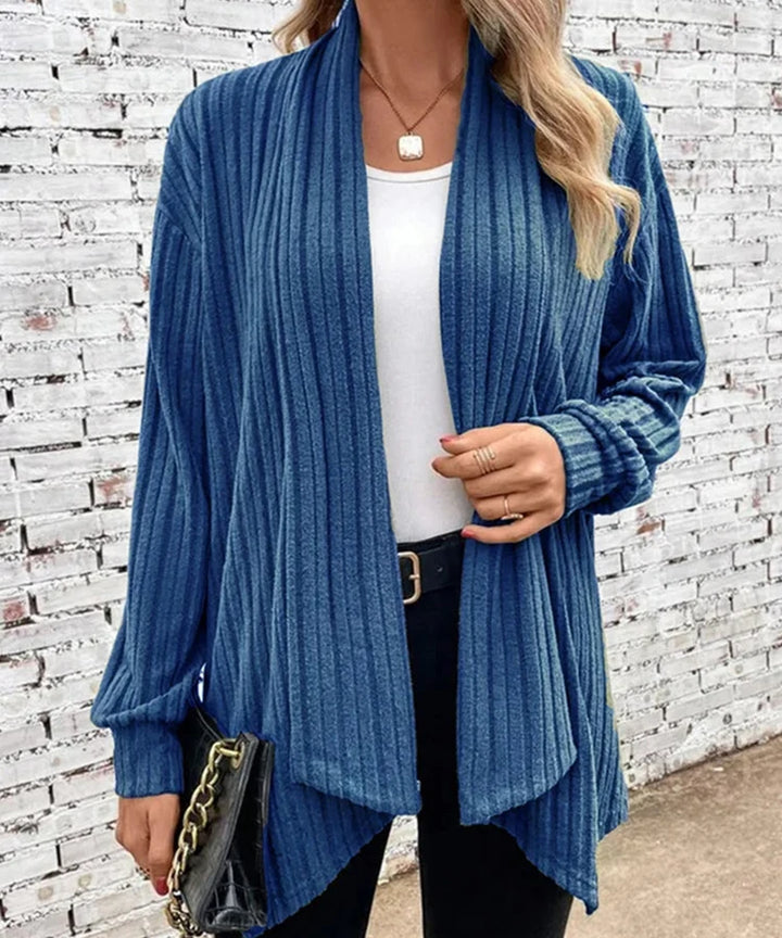 Woman wearing a blue ribbed cardigan over a white top and black pants, standing against a white brick wall.