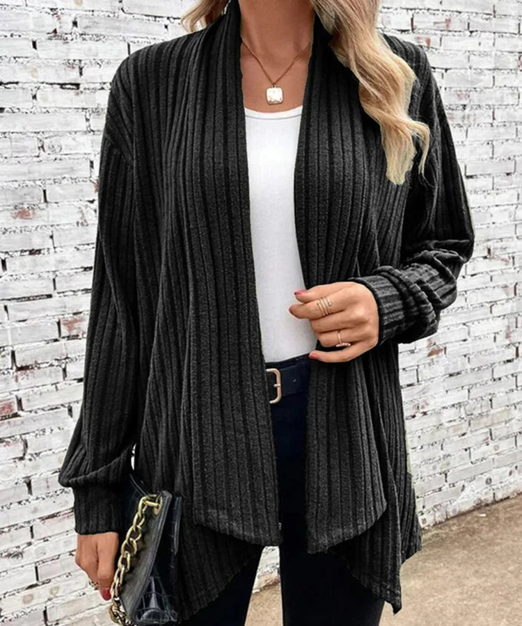 Person wearing a dark gray ribbed cardigan over a white top with a brick wall background