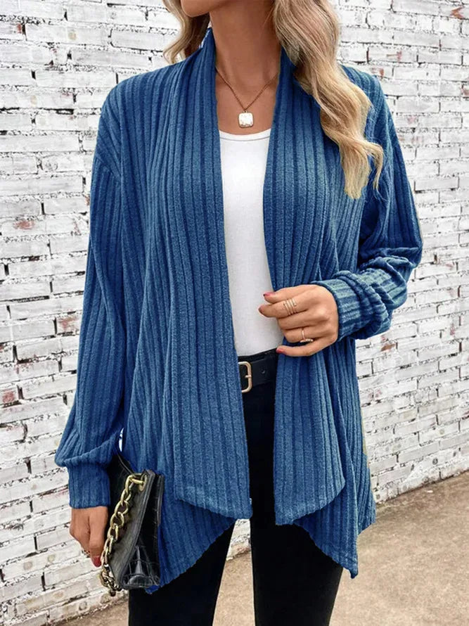 Person wearing a blue ribbed cardigan over a white top and black pants, standing against a white brick wall.