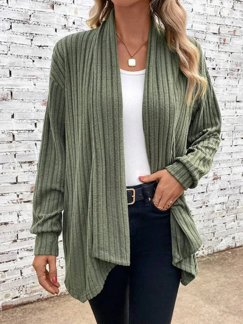 Woman wearing a green ribbed cardigan over a white top and dark jeans against a white brick wall.