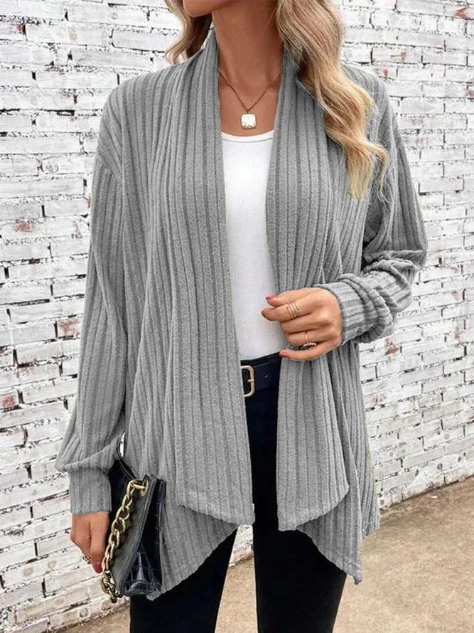 Woman wearing a gray ribbed cardigan over a white top and black pants, standing against a light brick wall.