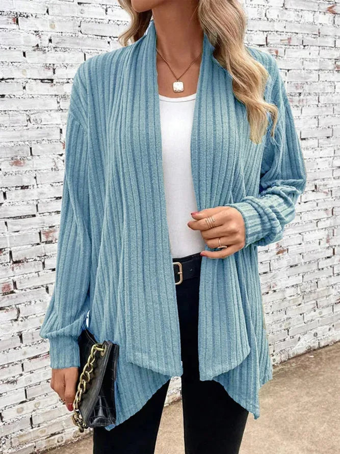 Woman wearing a light blue ribbed cardigan over a white top and black pants, standing against a white brick wall.