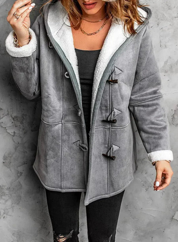 Person wearing a gray coat with white fur lining against a concrete wall.